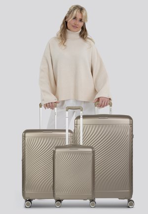 Trois valises en métal doré avec des motifs texturés ondulés, dotées de poignées télescopiques et de roues, placées devant une personne portant un pull léger.