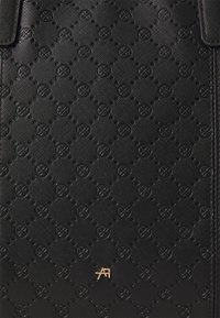 Surface texturée noire de sac avec des motifs circulaires en relief et un petit logo métallique doré près du centre en bas.