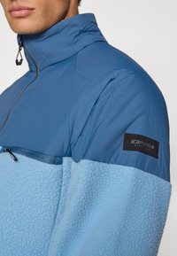 Pullover a mezza zip in blu bicolore. Presenta una parte superiore liscia e una parte inferiore pelosa, con una patch del logo sulla manica.