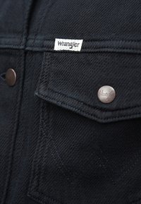 Crna traper jakna s Wrangler etiketom, bakrenim dugmadi i karakterističnim uzorcima šivanja na džepovima. Teksturalna tkanina.