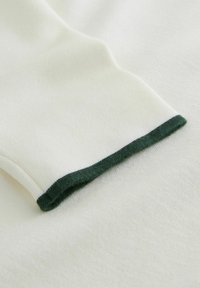 White fabric with a soft texture, featuring a dark green ribbed cuff. Seam detail visible, emphasizing the structure of the garment.