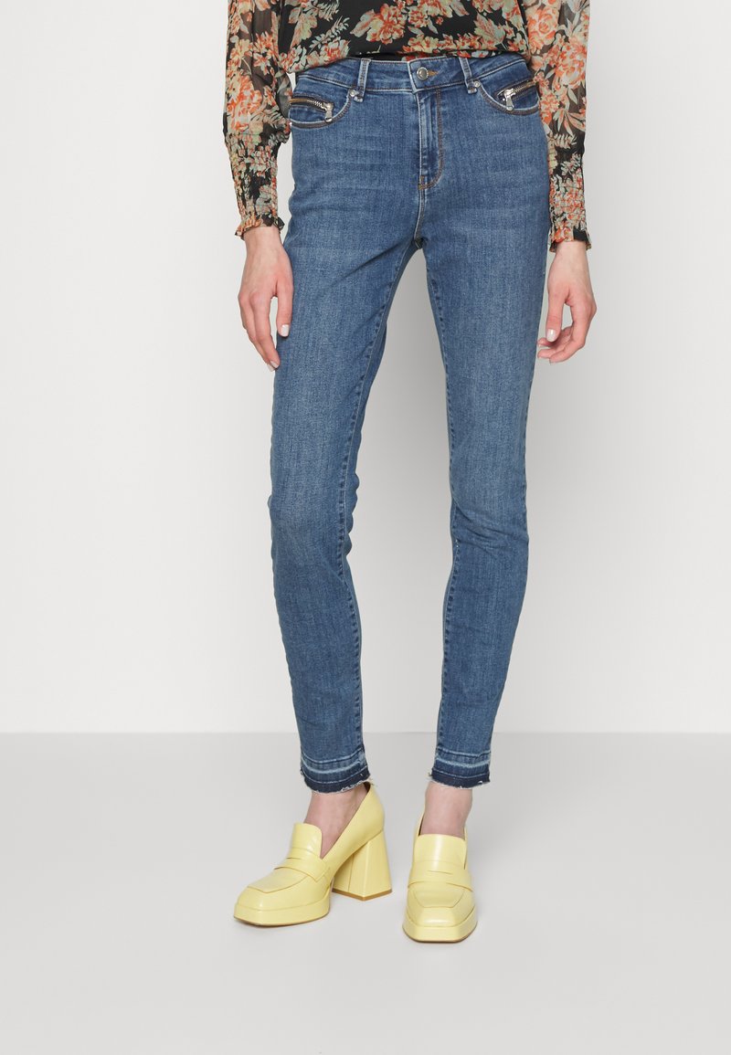 Jeans de denim azul skinny com cinco bolsos e bainha desgastada. Combinados com sapatos de salto bloco amarelos. Blusa de mangas longas com padrão floral.