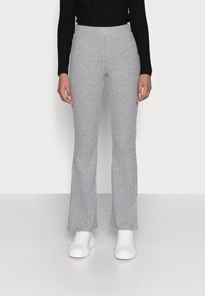 Vrouw die lichtgrijze geribbelde gebreide broek met hoge taille, zwart top met lange mouwen en witte sneakers draagt, staand op een effen witte vloer.