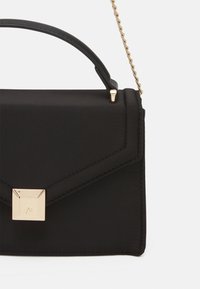 Sac à main noir avec une finition en tissu lisse, un design géométrique, des éléments métalliques dorés et une bandoulière chaîne amovible. Dispose d'une fermeture à boucle sécurisée.