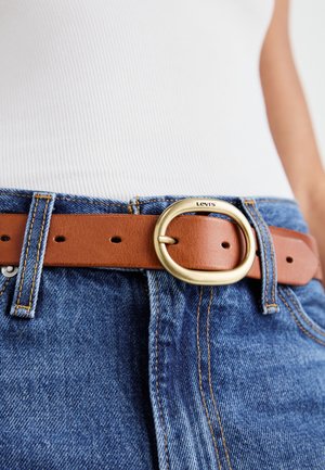 Brown leather belt with a large gold circular buckle, worn at the waist over blue denim. Stitched accents visible on the belt and pants.