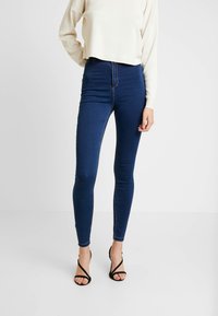 Jeans de mezclilla de cintura alta en azul oscuro, diseño ajustado con ligero estiramiento, que presentan un estilo clásico de cinco bolsillos y detalles de costura.