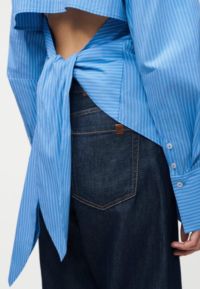 Hátsó nézet egy sötétkék farmernadrágot és kék csíkos inget viselő személyről, akinek az ingjét hátul alul megkötötték, így a derékvonal fölötti bőr látható.