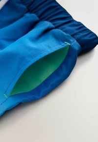 Sinised ujumispüksid rohelise sisemise tasku detailiga, millel on elastne vöökoht ja lamedad õmblused. Kangas näib olevat kerge ja sile.