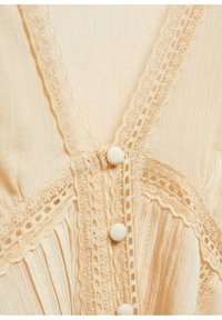 Top crema con scollo a V caratterizzato da un tessuto testurizzato, delicata rifinitura in pizzo e bottoni rivestiti in tessuto lungo la parte anteriore. Il design plissettato aggiunge profondità.
