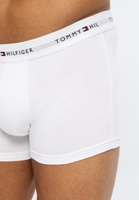 Κοντινό πλάνο ενός ατόμου που φοράει λευκά ανδρικά εσώρουχα Tommy Hilfiger με ζωνάρι με το λογότυπο, σε λευκό φόντο.