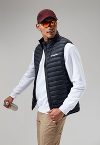 Zwarte puffer vest over een wit sweatshirt, beige broek en kastanjebruine pet. Model houdt een zilveren waterfles vast en draagt oranje getinte zonnebril.