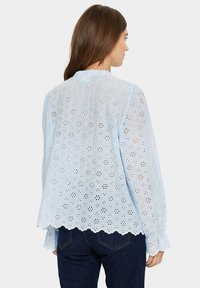 Blusa azzurro chiaro con motivi floreali ricamati, orlo a scallop e maniche lunghe con polsini con bottoni, realizzata in un materiale di cotone trasparente.