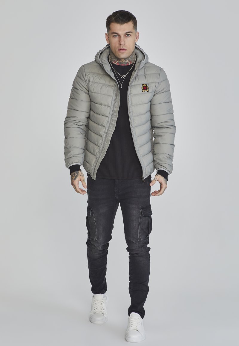 Chaqueta acolchada gris con capucha, que presenta un pequeño logo bordado. Llevada sobre un suéter negro de canalé y combinada con pantalones cargo negros y zapatillas blancas.