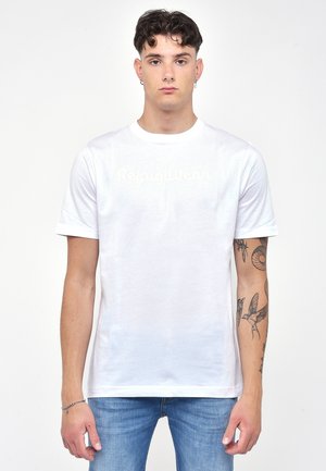 T-shirt bianco in cotone con maniche corte, scollatura tonda e logo "RefugilWear" in stampa chiara. Indossato con jeans blu, che mettono in mostra tatuaggi sulle braccia.