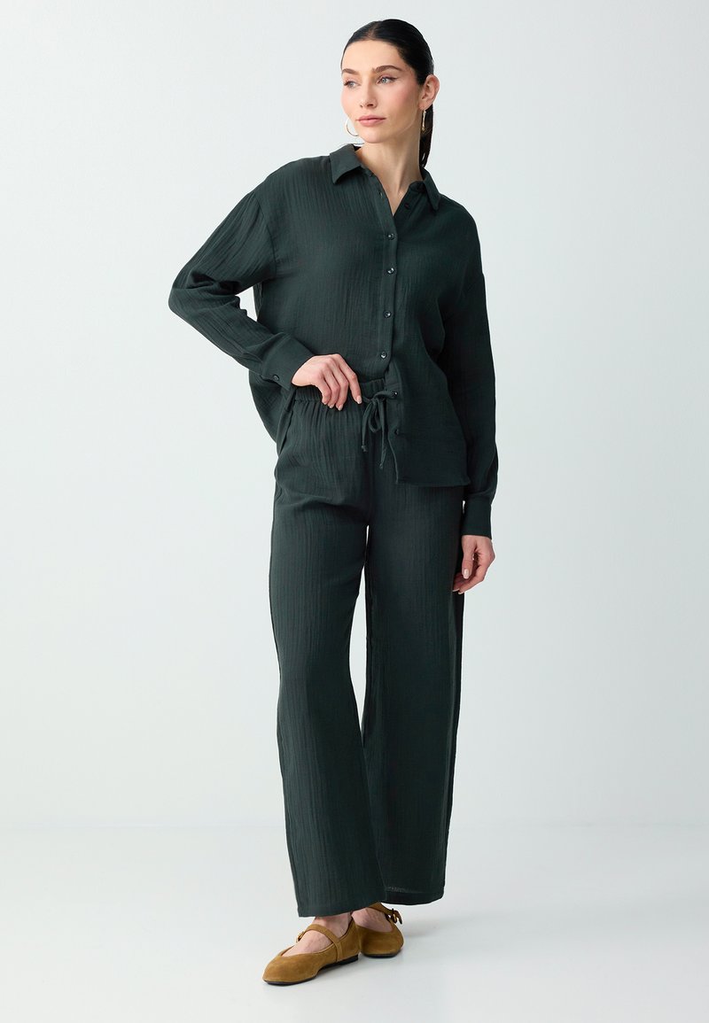 Femme aux cheveux foncés en queue de cheval basse, vêtue d'une chemise verte foncée à boutons et manches longues, et d’un pantalon large assorti avec des chaussures marron.