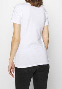 Vrouw in een eenvoudig wit T-shirt met korte mouwen en zwarte jeans, gezien vanuit de achterkant op een witte achtergrond.