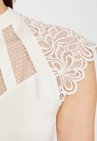 Vêtement blanc avec des accents en dentelle délicate sur les épaules, présentant un panneau en maille et une texture de tissu lisse, mettant en valeur des motifs floraux.