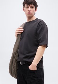 Νεαρός άνδρας που φοράει μαύρο t-shirt και παντελόνι, κρατώντας ένα χακί μπουφάν πάνω στον έναν ώμο, μπροστά σε μια απλή φωτεινή επιφάνεια.