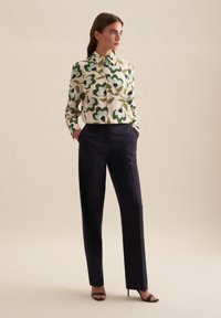 Camicia a maniche lunghe con motivo floreale verde e blu, abbinata a pantaloni neri su misura e tacchi neri, su uno sfondo neutro.