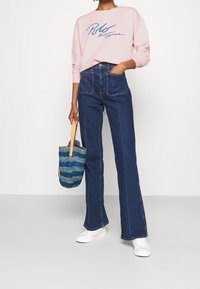 Blå denim med utsvängda byxor med framfickor, matched med en ljusrosa sweatshirt med en marinblå logotyp, och en vävd blå strandväska. Vita sneakers.