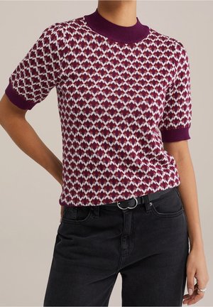 Pull à manches courtes présentant un motif losange en bordeaux et blanc, avec un col ras du cou et des poignets côtelés bordeaux. Porté avec un pantalon noir.