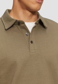 Polo en khaki avec un col, fabriqué dans un tissu texturé, deux boutons noirs, et un sous-vêtement blanc superposé visible en dessous.