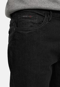 Nærbilde av lomme på svarte denimjeans med Indicode-merke, brukt med en grå topp.