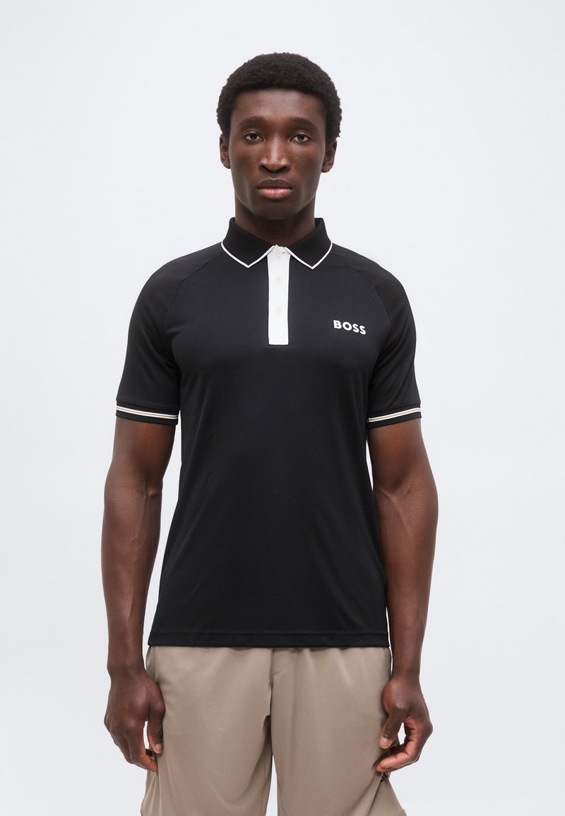 Νέος άνδρας με μαύρο πόλο μπλουζάκι με λευκή επένδυση και λογότυπο «BOSS», συνδυασμένο με μπεζ παντελόνια, στέκεται μπροστά σε απλό φόντο.