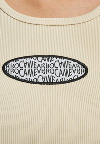 Beige ribbet top med en rund hvid lap med sort tekst, der gentager "Brocamewear", og en sort broderet kant.