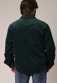 Dunkelgrünes Cordhemd mit sichtbarer Ripptextur, spitzem Kragen und geknöpften Manschetten, kombiniert mit blauen Jeans.