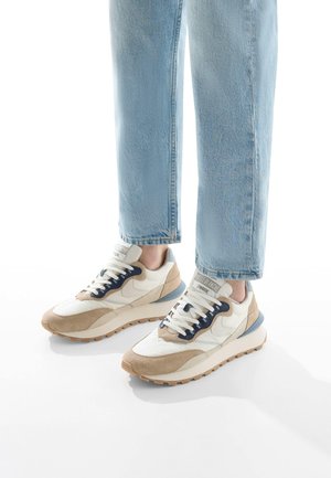 Sneaker mit einem weißen Stoff-Obermaterial, beigen Wildleder-Elementen, dunkelblauen Schnürsenkeln und einer strukturierten Gummisohle. Helle Jeans-Hosen sind teilweise sichtbar.