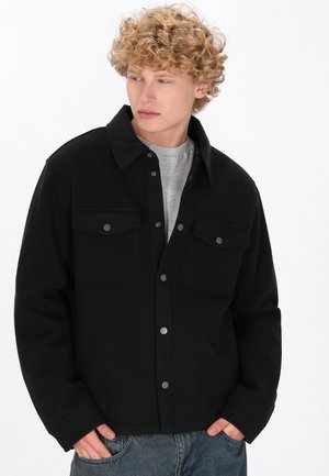 Black button-up jacket made of thick fabric, featuring two chest pockets, metal snap buttons, and a collared neckline.