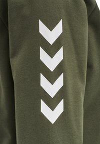Sweatshirt vert olive avec une texture douce, présentant trois grands motifs en chevron blancs alignés verticalement sur la manche.