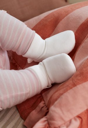 Gambe di un bambino con pantaloni a righe rosa e bianche che indossano morbidi scarpette bianche, poggiate su una superficie di tessuto a righe rosse e rosa.