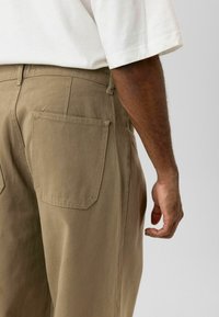 Beige, loszittende broek met dubbele achterzakken en een gestructureerde stof, gedragen met een wit shirt met korte mouwen.