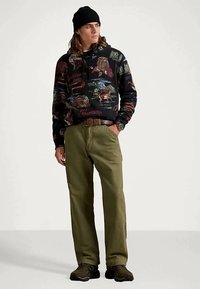 Jonge man draagt een zwarte beanie, een zwart hoodie met wildlife afbeeldingen, olijfgroene broek, een bruine riem en donkere sneakers, terwijl hij met zijn hand in zijn zak staat.