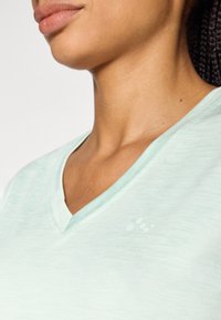 Ανοιχτό πράσινο πουκάμισο με V λαιμόκοψη, με απαλό υφή και διακριτική λεπτομέρεια λογότυπου στο στήθος. Το ύφασμα φαίνεται διαπνέον και ελαφρύ.