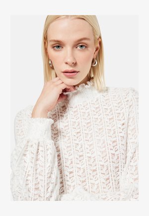 Donna bionda con occhi azzurri indossa orecchini a cerchio d'argento e una camicetta bianca in pizzo con collo alto, riposando delicatamente la mano vicino al mento.