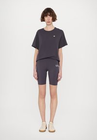 Tricou din bumbac gri cu mâneci scurte, combinat cu pantaloni scurți pentru bicicletă asortați care au un imprimeu cu logo. Poartă adidași ușori.