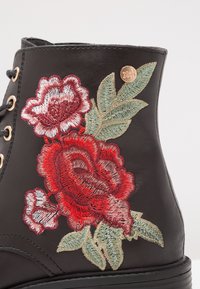 Bottine en cuir noir avec broderie florale rouge et blanche, agrémentée de feuilles vertes et d'un petit accent doré. Design à lacets.