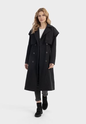 Kvinde med blondt hår iført en lang sort dobbeltradet frakke, grå opkrøjede jeans og sorte snørestøvler, går fremad.
