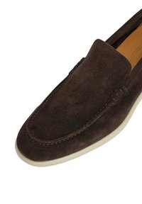 Zapato marrón de ante tipo slip-on con punta redondeada, detalles de costura y suela de goma de color claro. Textura suave y acentos mínimos.