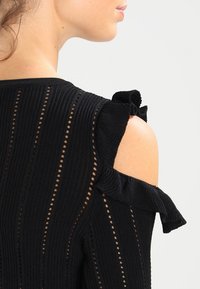 T-shirt en tricot noir avec des épaules dénudées, texture côtelée et accents volants sur les manches ; comprend un motif à rayures verticales avec des découpes.