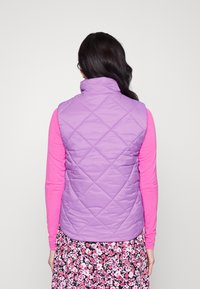 Gilet matelassé violet clair avec un col haut, associé à une chemise à manches longues rose, sur fond d'une jupe à motifs floraux.