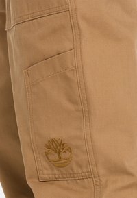 Khaki cargo pants featuring a side pocket and an embroidered logo. The fabric is durable with a textured finish, ideal for outdoor wear.
