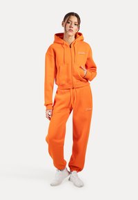 Haut à zip orange avec poches avant et pantalon de survêtement assorti. Le tissu est doux. Comprend des poignets et une taille élastiques. Accents de logo blancs.