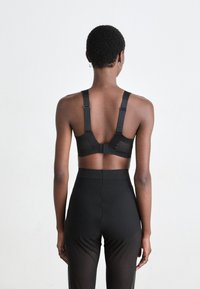 Persona con cabello corto que lleva un sujetador negro y leggings negros transparentes de cintura alta, de frente a un fondo blanco liso.