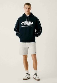 Felpa navy con logo e testo bianchi, con lacci bianchi e cuciture a contrasto. Abbinata a pantaloni corte grigi chiari e sneaker nere.