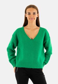 Maglione verde a maglia con scollo a V, vestibilità morbida e orlo a coste. Texture morbida e maniche lunghe, abbinato a pantaloni neri.