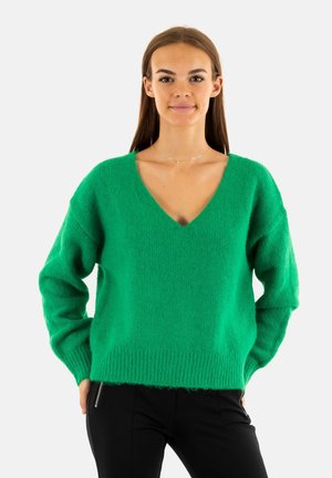 Un pull en maille verte avec un col en V, coupe lâche et ourlet côtelé. Texture douce et manches longues, associé à un pantalon noir.